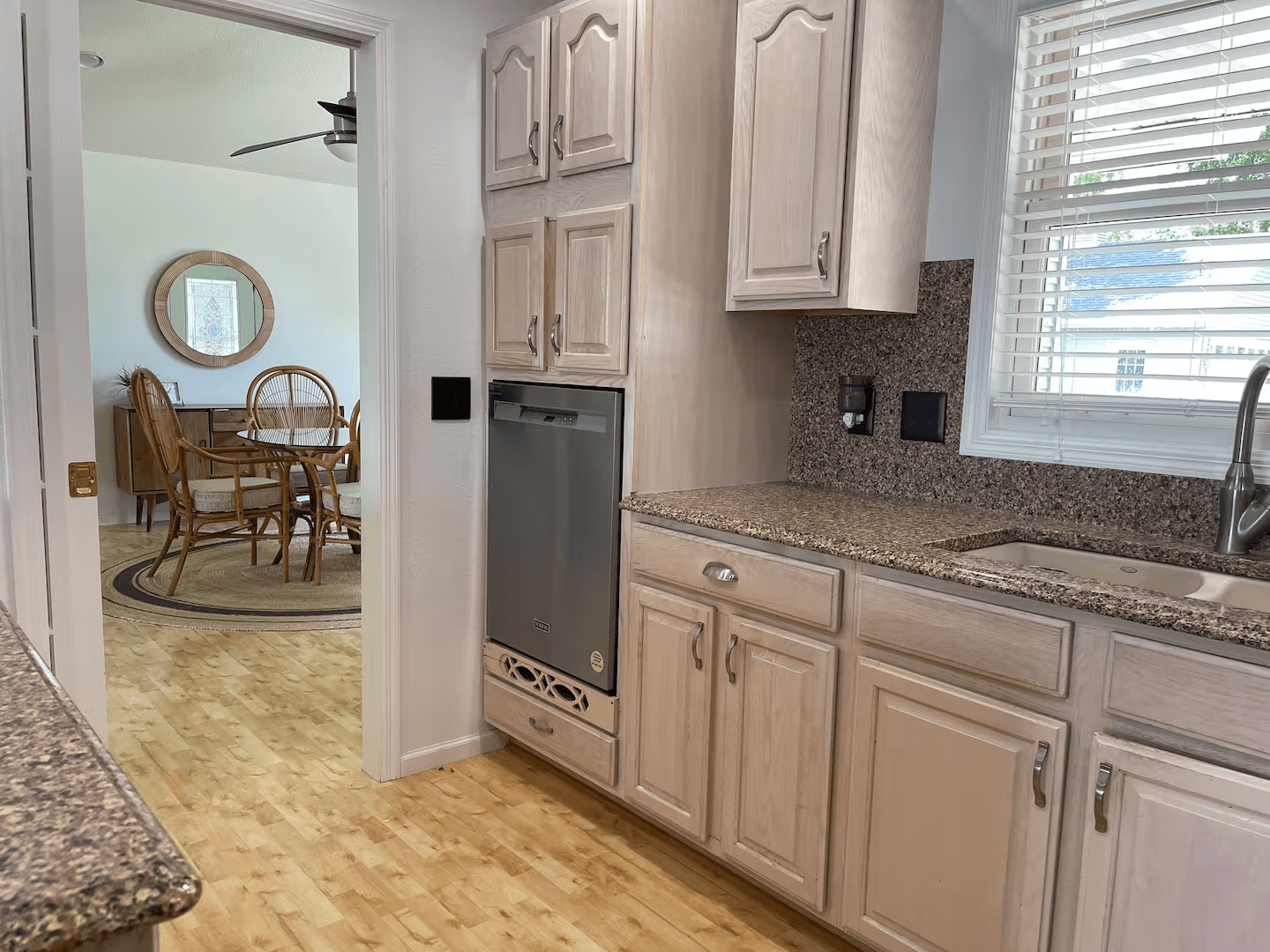 A contemporary kitchen with light wood cabinetry and sink with window above. The dining area is visible through an open doorway.