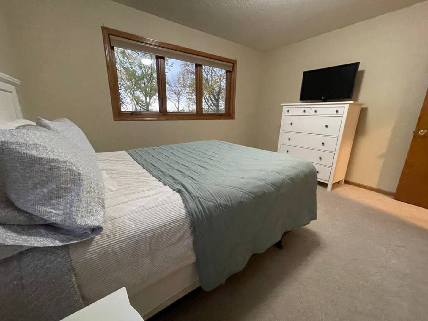 A bedroom with a bed facing a white dresser and a flat-screen TV with sunlight coming from three-pane window with blinds.
