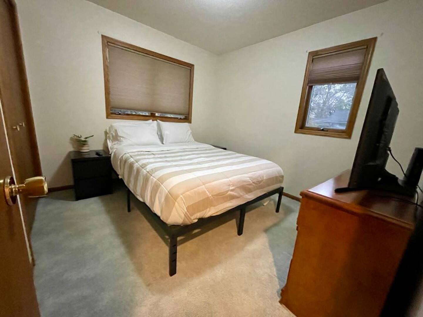 A bedroom with a bed facing a dresser with a flat-screen TV on top; two windows with blinds allow for natural light.