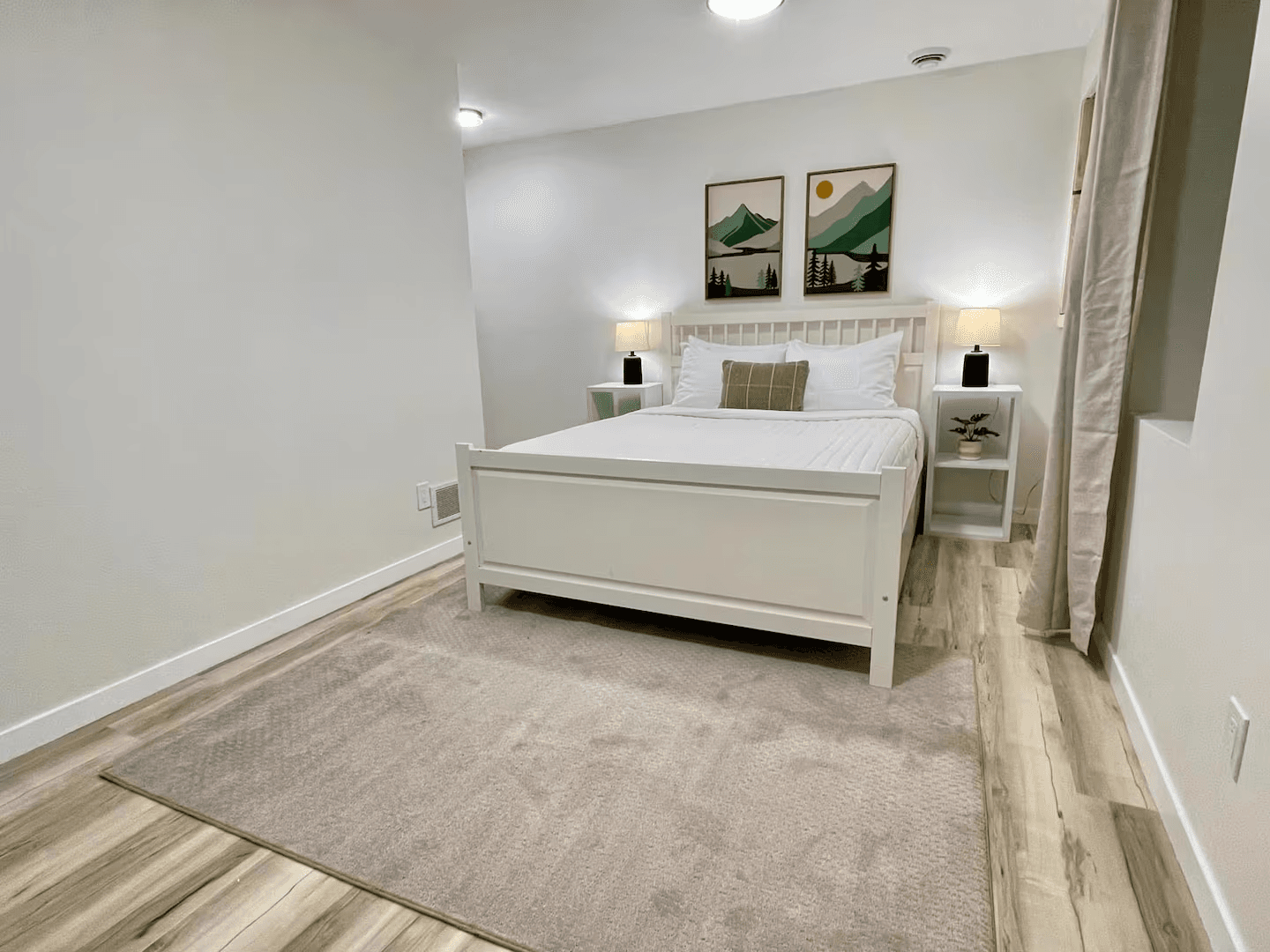 A cozy bedroom with light wood flooring and a large area rug. A bed with a white painted wooden frame stands in the center flanked by matching nightstands topped with table lamps.