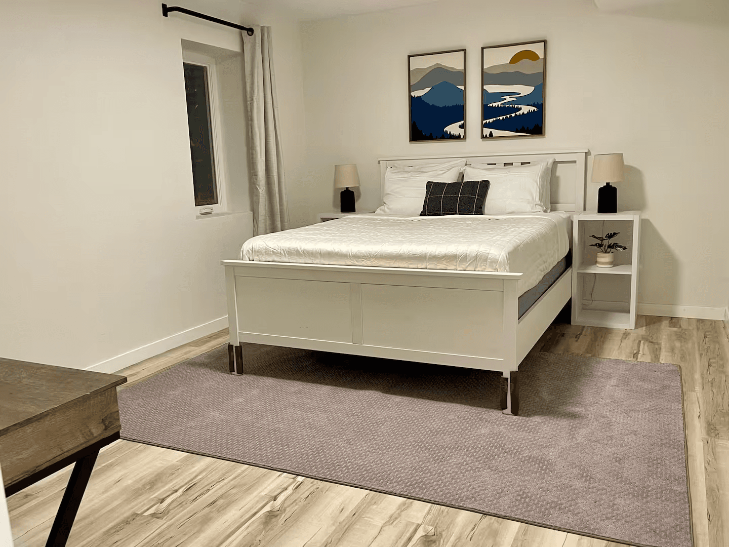 A cozy bedroom with light wood flooring and a large area rug. A bed with a white painted wooden frame stands in the center flanked by matching nightstands topped with table lamps.
