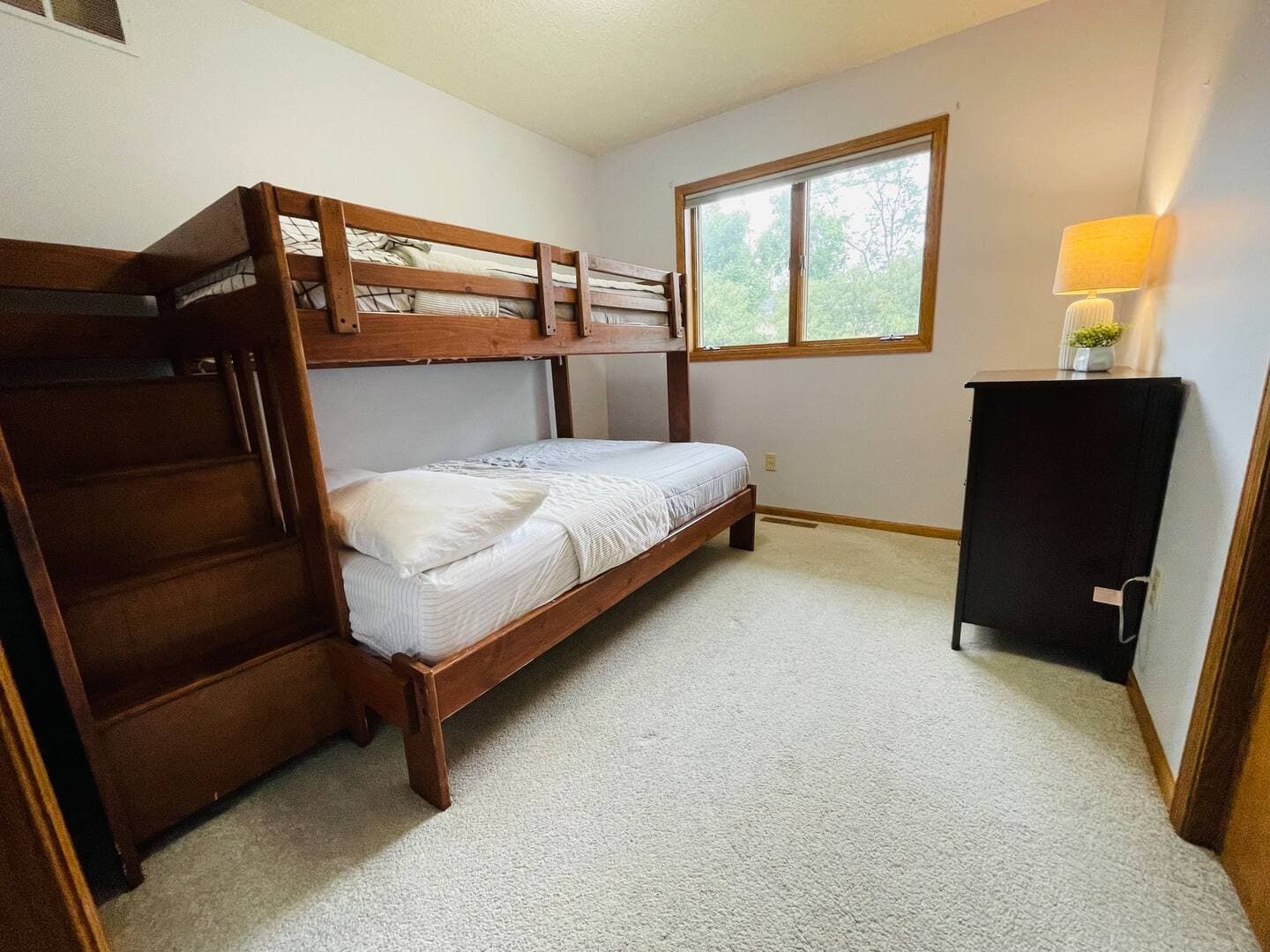 A cozy bedroom with light wood flooring features a bunk bed with white bedding. A large window overlooks trees.