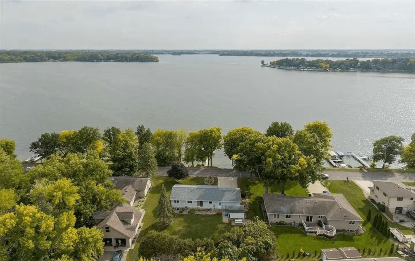 An aerial view shows a residential neighborhood with several houses, lush green trees, and Madison Lake in the background.