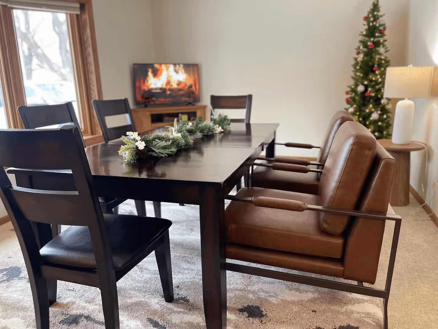 A spacious, light-filled dining area featuring a rectangular wooden table, six chairs with dark cushions, and a large patterned area rug.