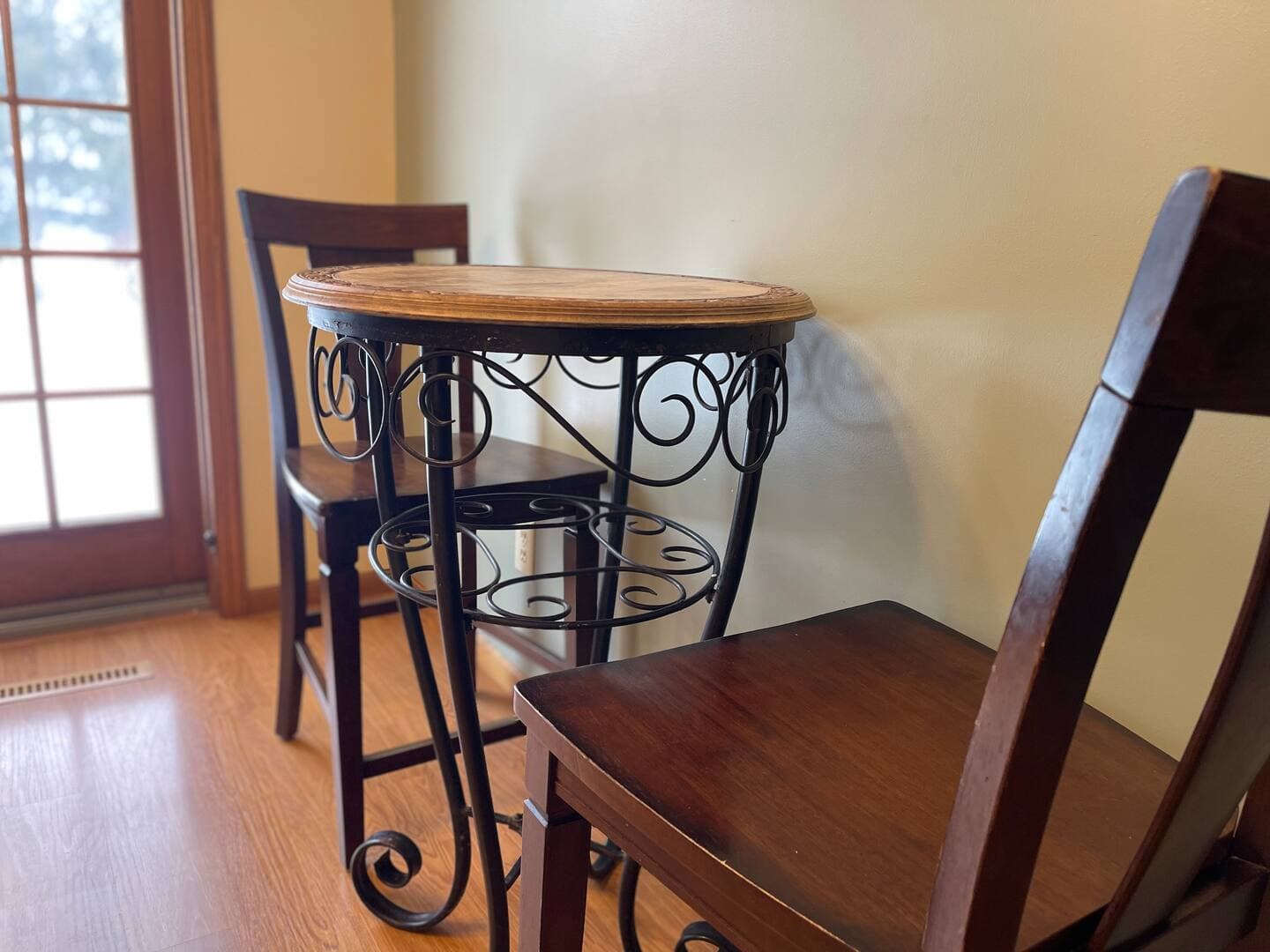 A cozy kitchen corner features a small round wooden table with an ornate metal base and two wooden chairs.