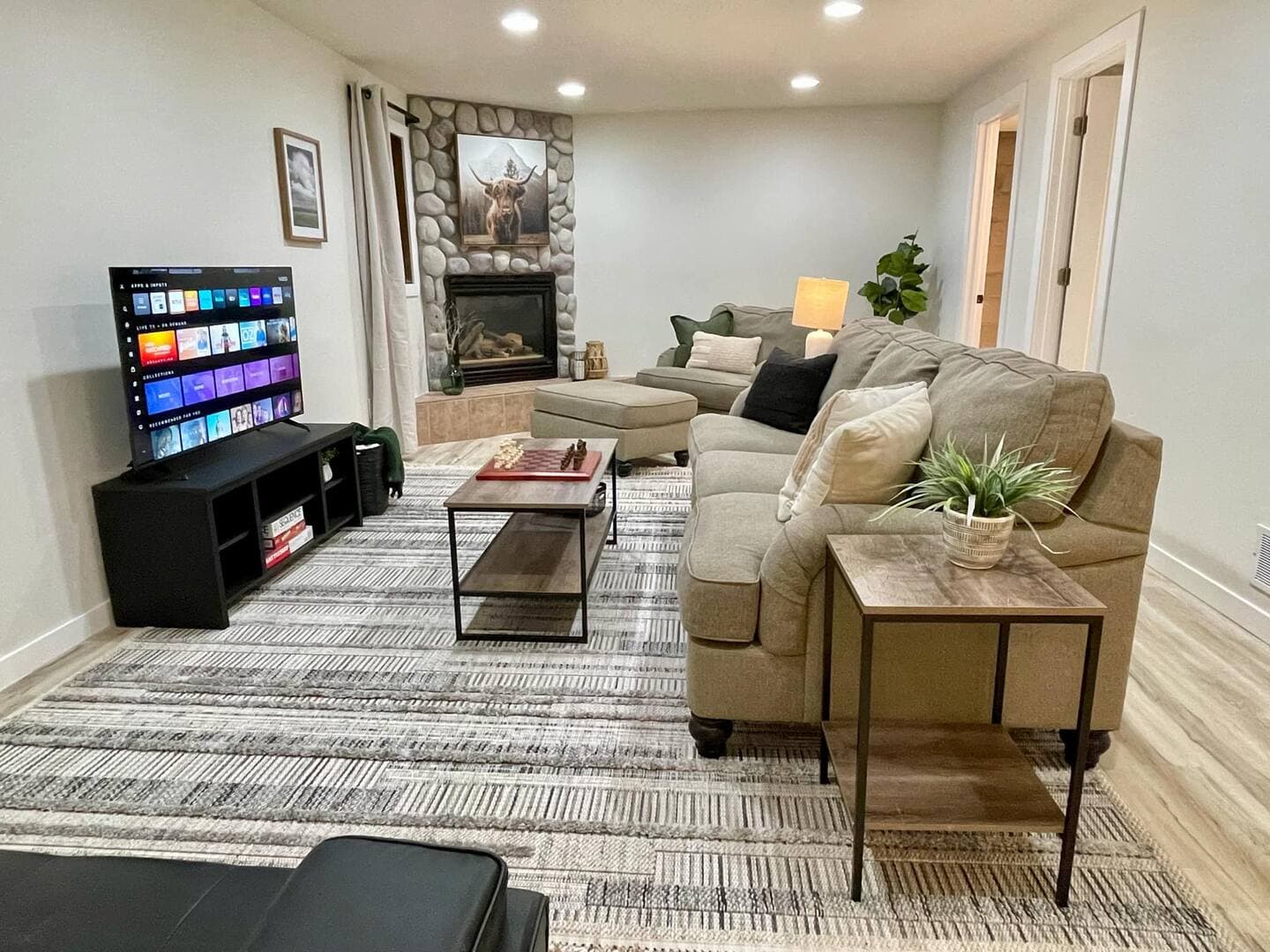 A cozy living room features a stone fireplace, a large TV, a sofa, a loveseat, and an armchair - all in tranquil earth tones.