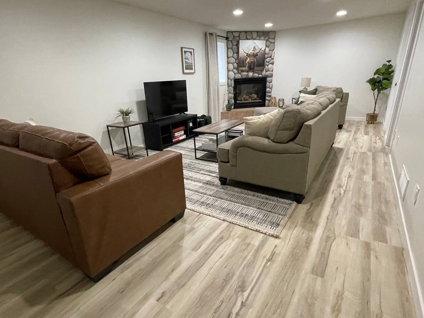 A cozy living room features a stone fireplace, a large TV, a sofa, a loveseat, and an armchair - all in tranquil earth tones.