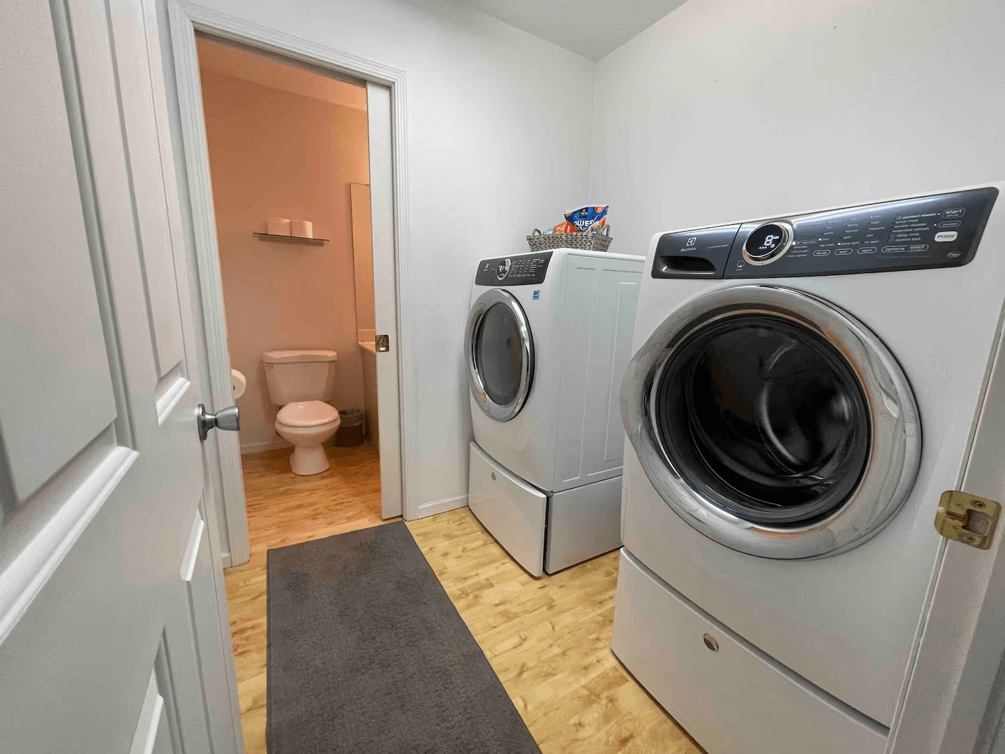 Laundry room with large washer and dryer and light wood floor. A bathroom is visible through an open doorway.