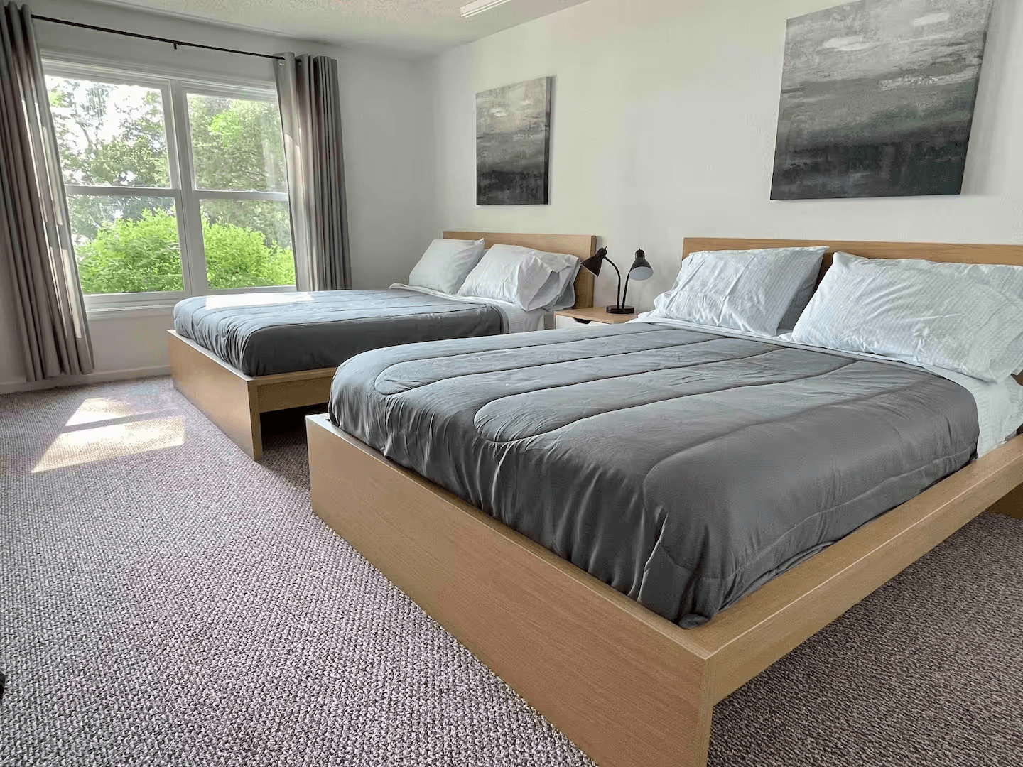 Bedroom with two queen beds on light wooden frames, wall-to-wall carpeting, and a large window with lots of natural light.