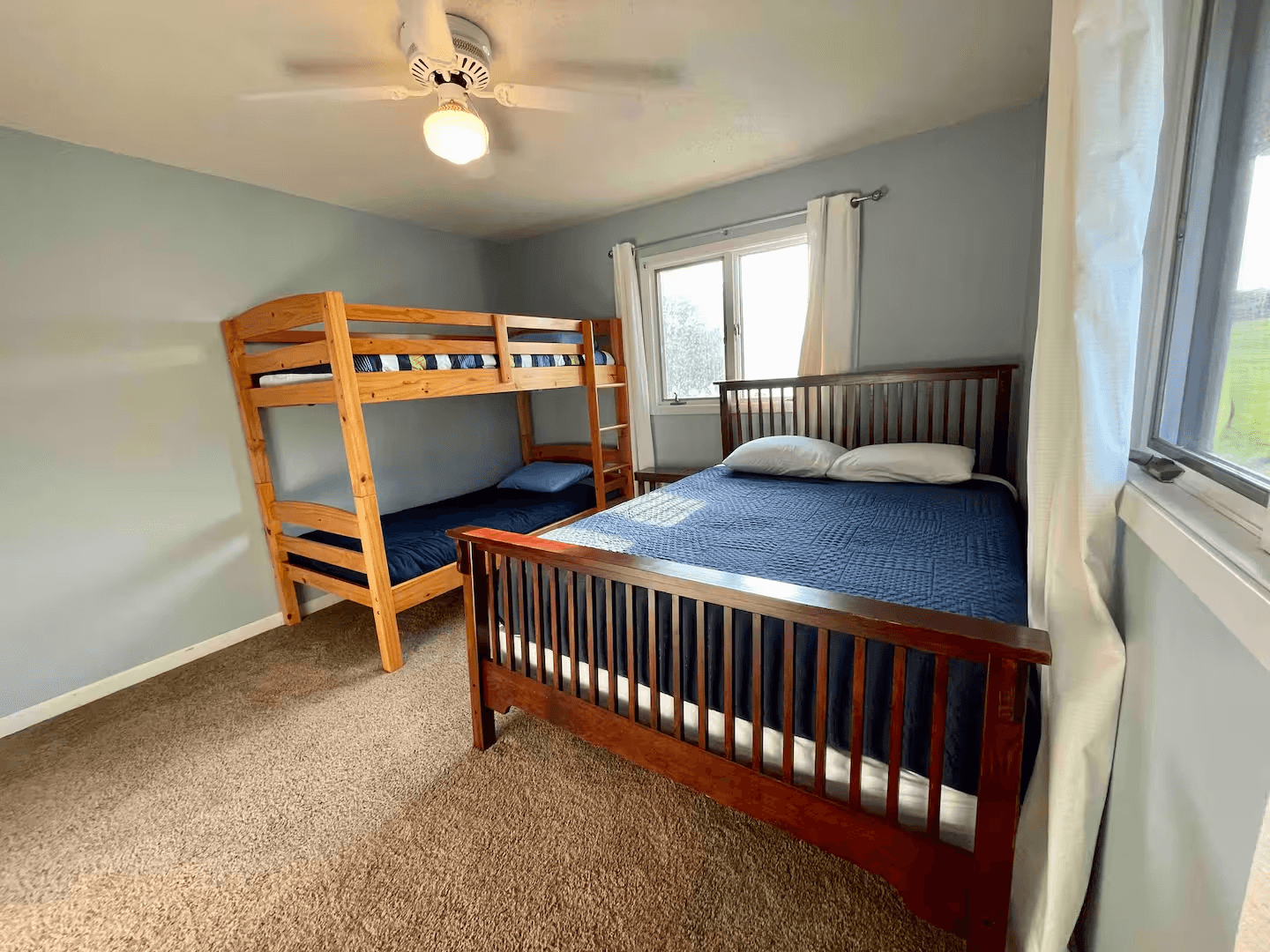 A carpeted bedroom with a large bed and a set of bunk beds. Two curtained windows let in plenty of natural light.