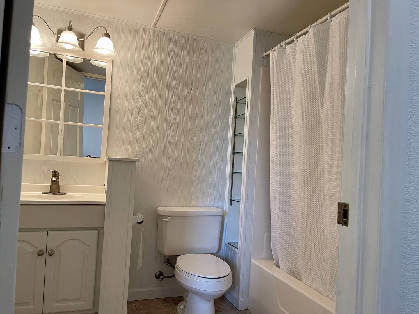 A clean, white bathroom with shower, toilet, and sink with vanity, mirror, and overhead lights.