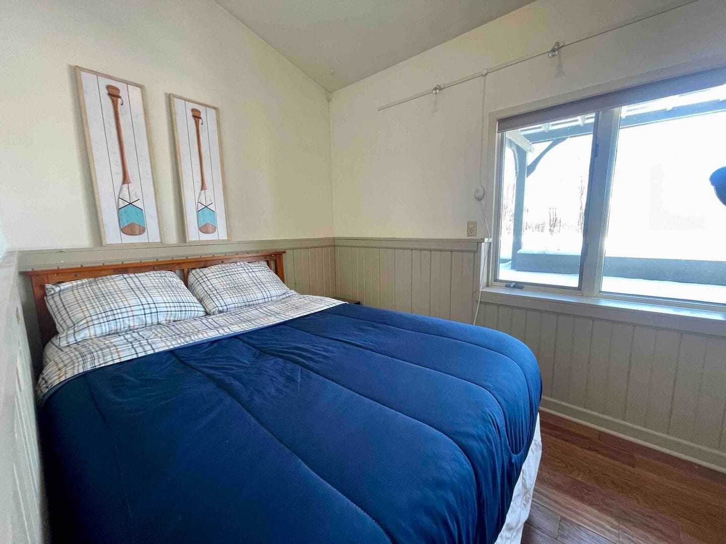 Bedroom with a cozy queen bed, light wood floors, and natural light from a large window.