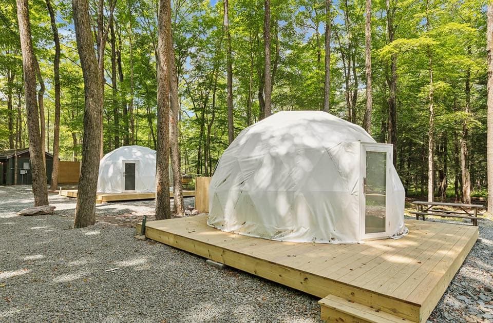 Two white geodesic domes on wooden platforms surrounded by trees in a gravel setting.